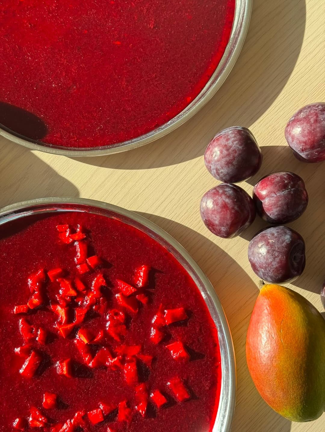 Overhead view of vibrant red fruit puree in metal trays next to fresh plums and a mango